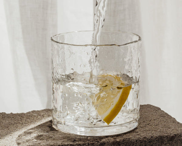 Clear glass being filled with water from a pitcher, with a lemon and ice cube inside.